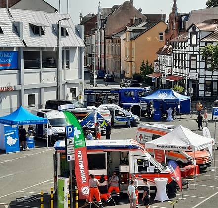 Blick auf einen Platz mit mehreren Einsatzfahrzeugen, darunter ein rotes Kreuzfahrzeug und Polizeiwagen. Zelte und Stände sind aufgestellt.