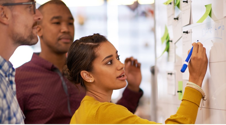 Drei Personen stehen vor einer Wand mit Notizen. Eine Person schreibt mit einem blauen Marker auf ein Blatt.
