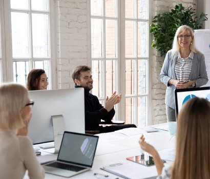 Frau steht vor Gruppe von vier Personen in einem hellen Raum mit großen Fenstern, Beratung zu Weiterbildung, Personen sitzen an Tisch mit Laptop und Tablet.